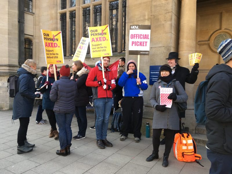 Some picketers (about 10) are photographed holding placards outside Exams Schools on Oxford High Street. Text on signs includes 'Official Picket' and 'Our Pension Axed'