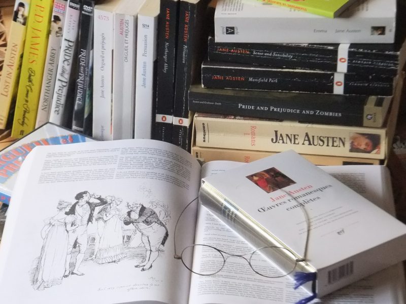 A Jane Austen novel is open on a table, with a pile of Austen novels in the background