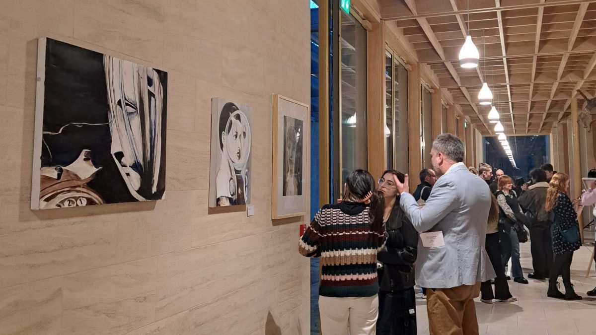 A photo of Worcester College's Exhibition 002. Three paintings are visible to the left of the image. Three people stand discussing the third painting, with a larger crowd visible behind them.
