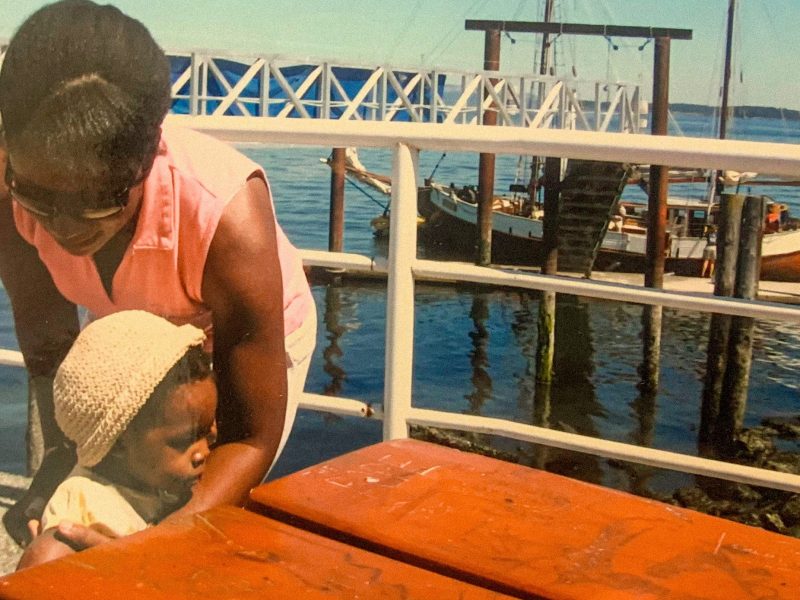 Childhood photo of Renée Orleans-Lindsay by the water with a boat on the water