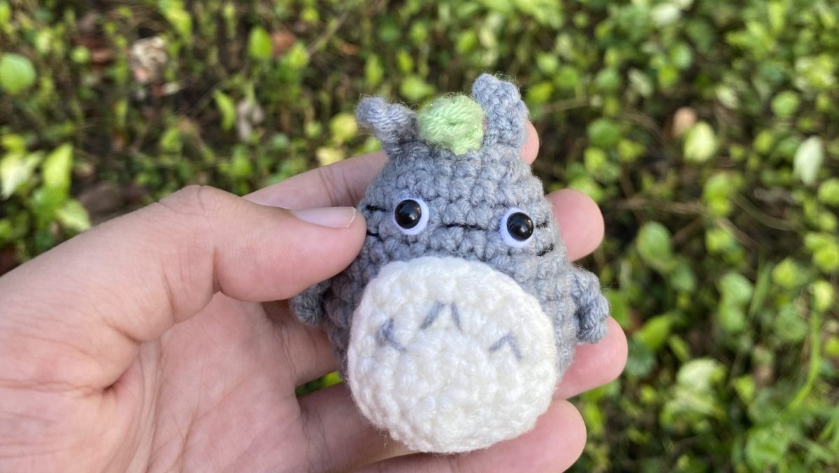Hand holding a small crocheted Totoro against a green background.