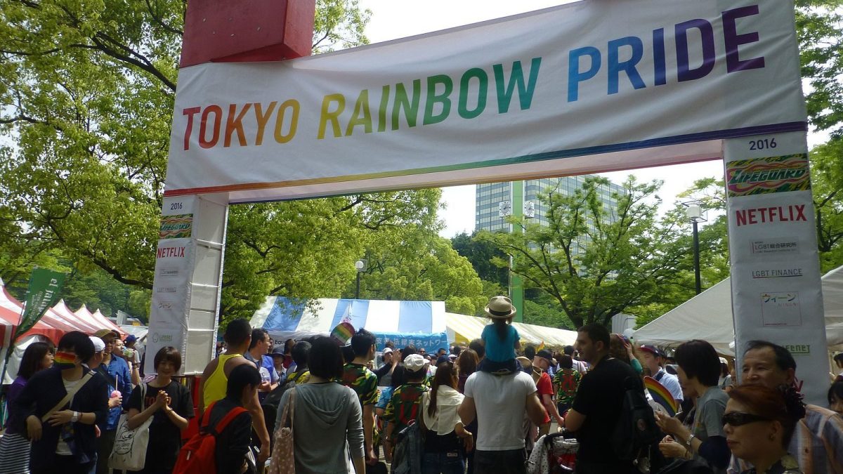 Crowds of people are gathered in a public park; a large banner overhead reads "Tokyo Rainbow Pride" in rainbow-colored letters.