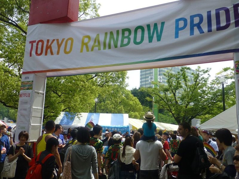 Crowds of people are gathered in a public park; a large banner overhead reads "Tokyo Rainbow Pride" in rainbow-colored letters.
