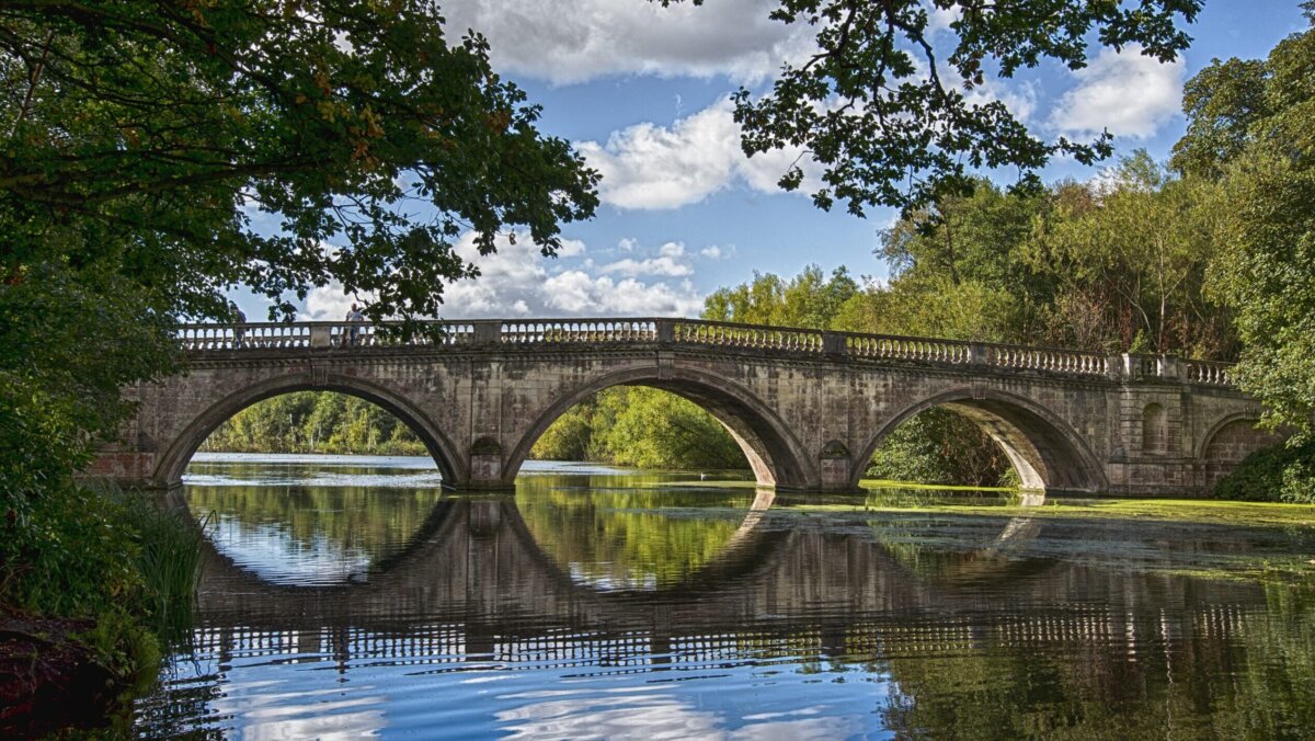 A photograph of a bridge across a lake. The bridge is old and has three arches, which are reflected on the surface of the water. It is flanked by trees on both sides.
