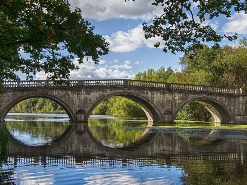 A photograph of a bridge across a lake. The bridge is old and has three arches, which are reflected on the surface of the water. It is flanked by trees on both sides.