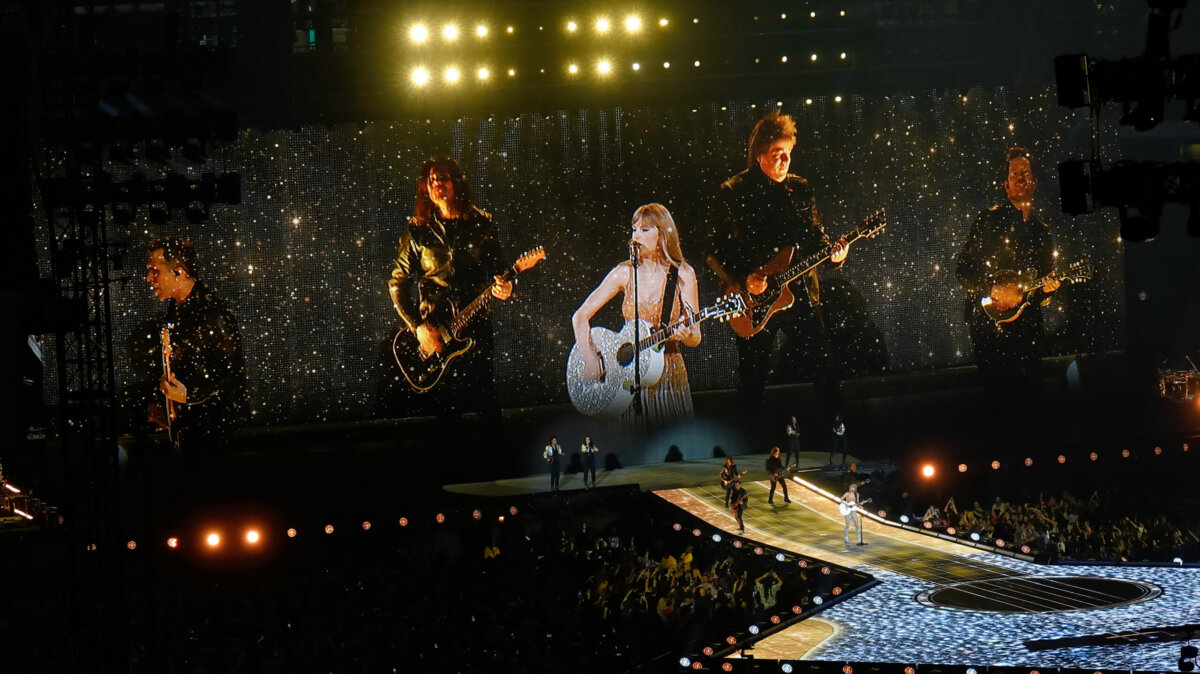 A photo taken at the Taylor Swift: The Eras Tour concert in Arlington. Taylor is on stage alongside various members of her band and backup singers. She is playing the guitar.