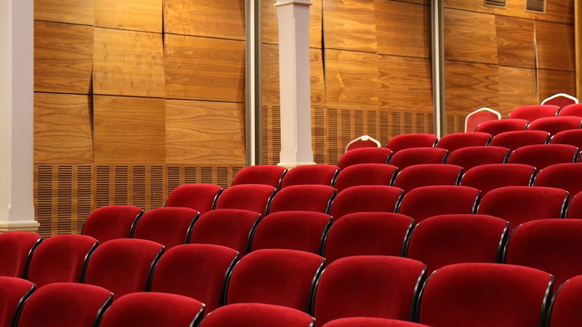 Picture of a theatre with wooden panelled walls, Roman style columns, and classic red velvet chairs.