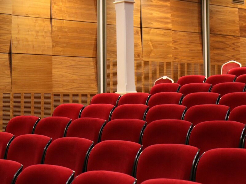 Picture of a theatre with wooden panelled walls, Roman style columns, and classic red velvet chairs.