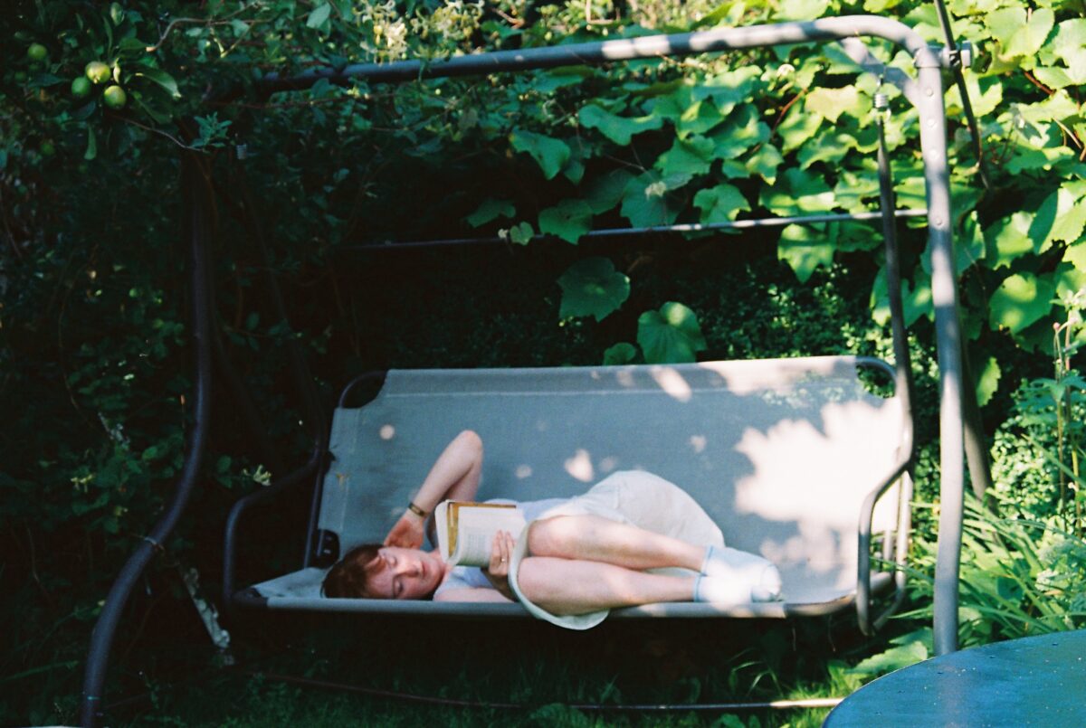 The photograph shows a girl in white lying on a garden swing chair reading a book. She is surrounded by the greenery of the over-hanging tree, bushes and grass.