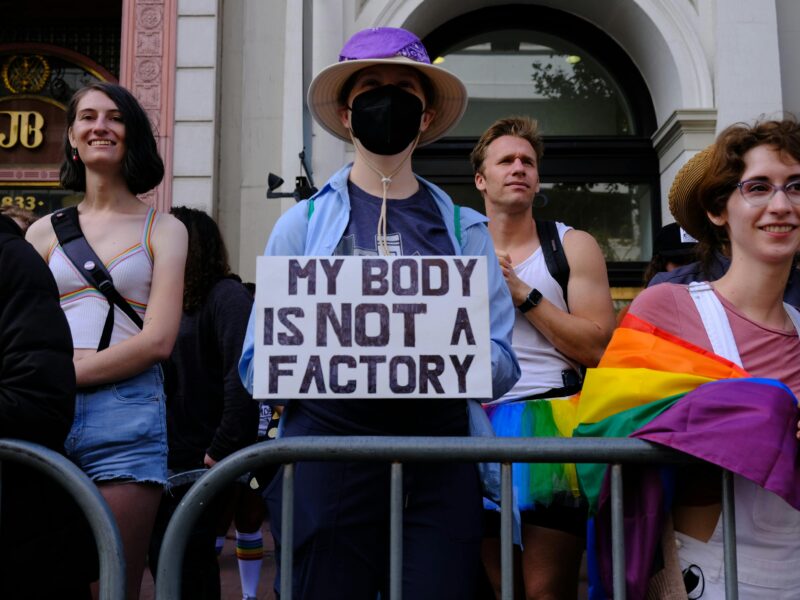 Image shows protesters. One holds a sign that reads 'my body is not a factory'.