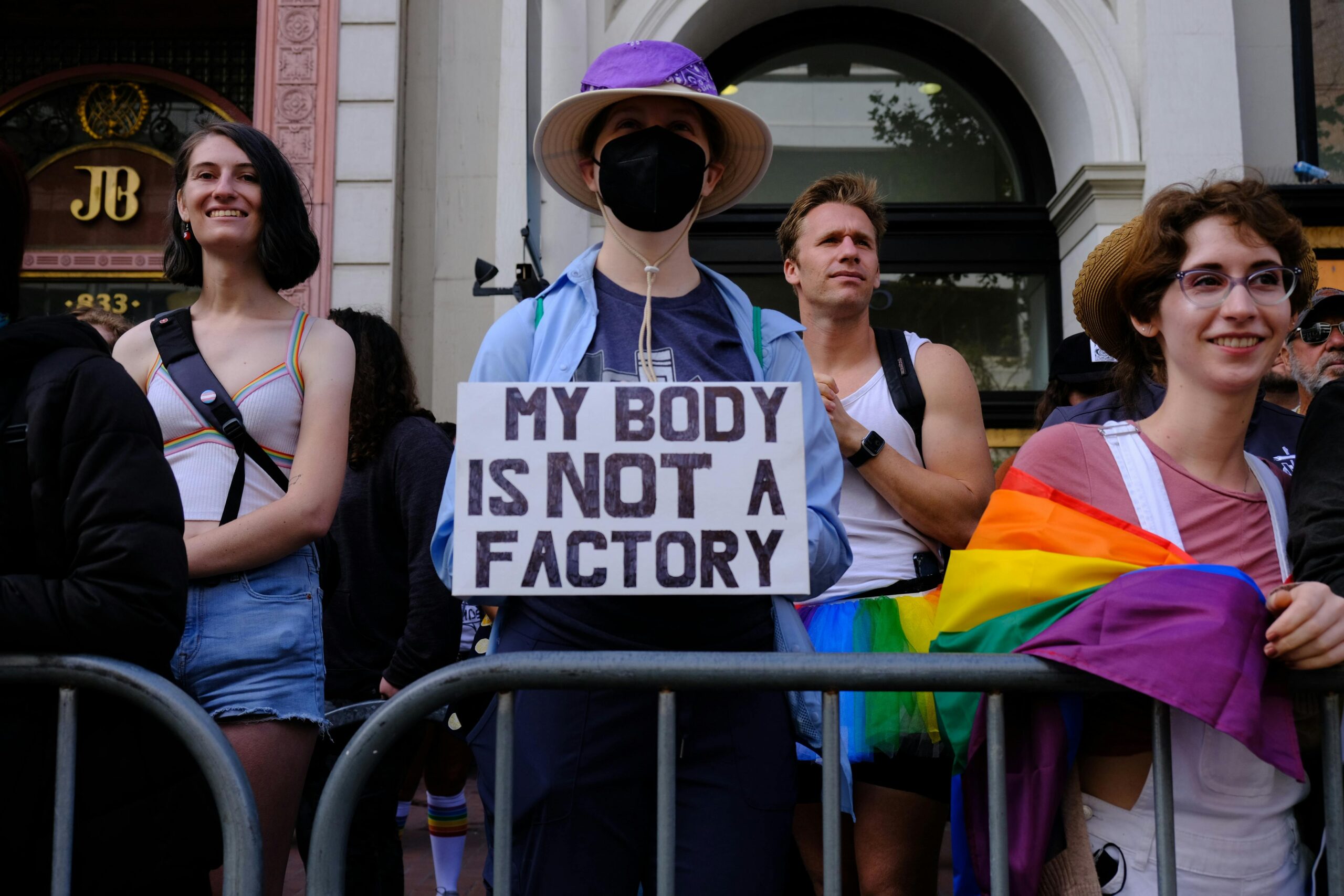 Image shows protesters. One holds a sign that reads 'my body is not a factory'.