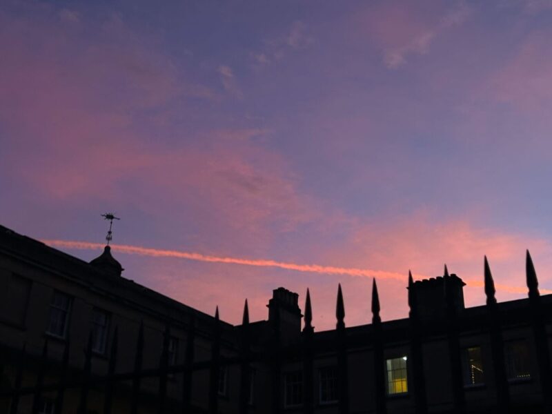 The photo is taken from a college quadrangle at sunset. It shows the top of College buildings in shadow. Behind them is a pink and blue sunset.
