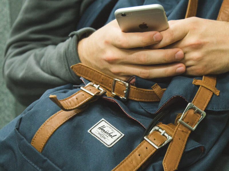 A man is holding a smartphone and a backpack whilst sitting in a chair.