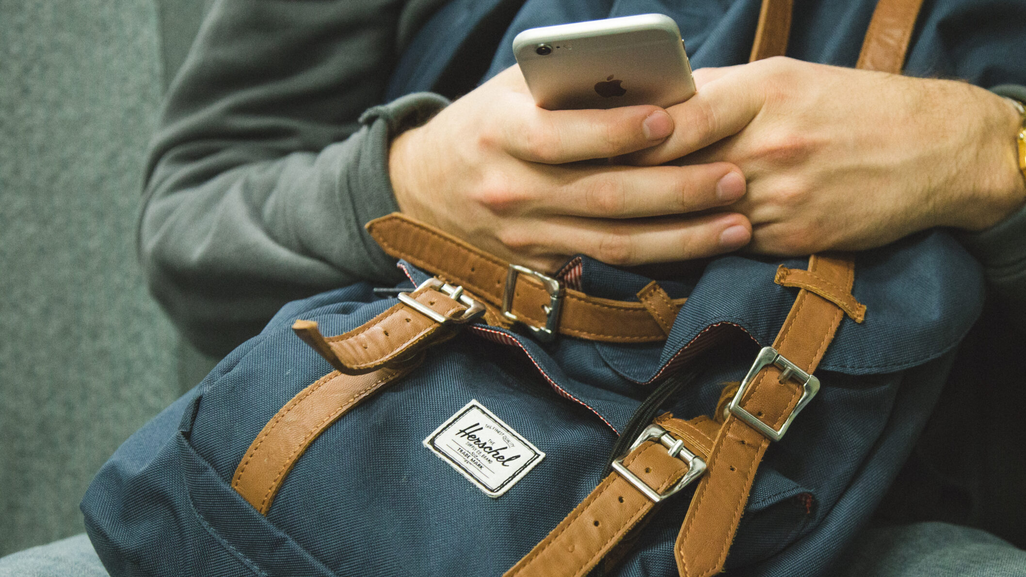 A man is holding a smartphone and a backpack whilst sitting in a chair.