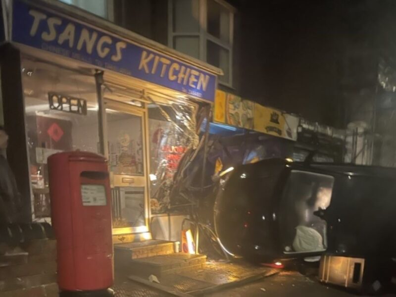 The image shows a car on its side, crashed into a shop. It is dark and there are yellow lights. It is a photo of the crash described in the article.