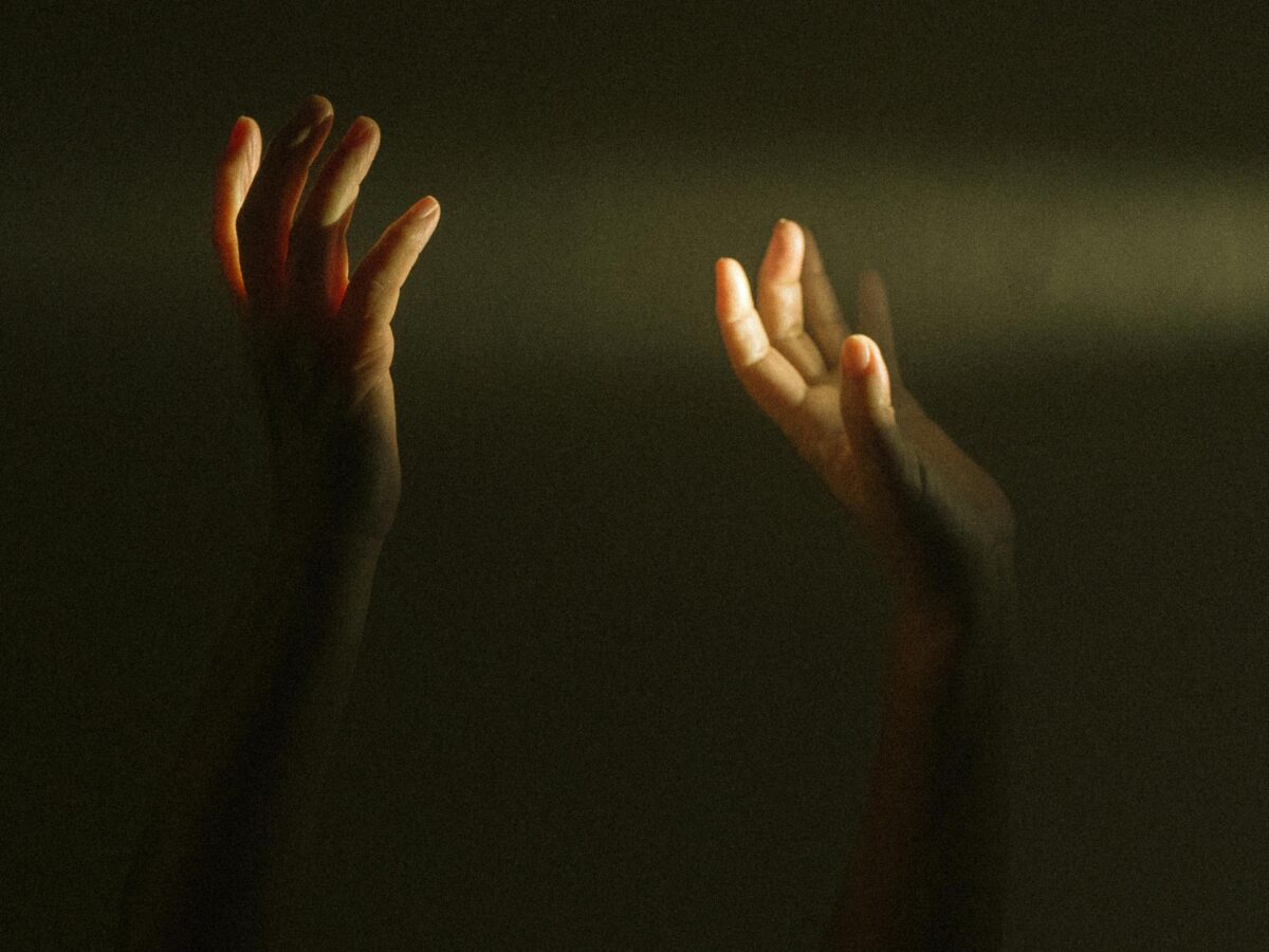 Photo shows two hands reaching up, against a dark background with one ray of light going horizontally across the frame.