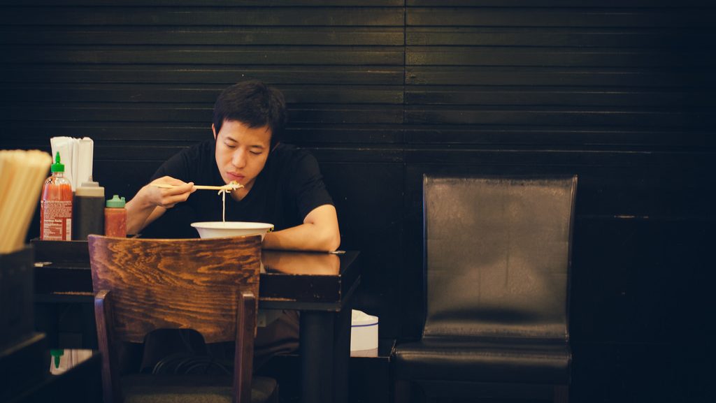 A man sits at a table alone, eating a bowl of ramen.