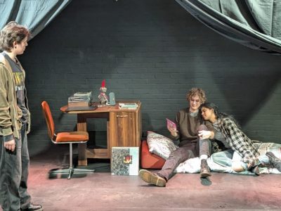 A photo of three cast members on set. Two are lying down whilst one is standing and speaking to them.