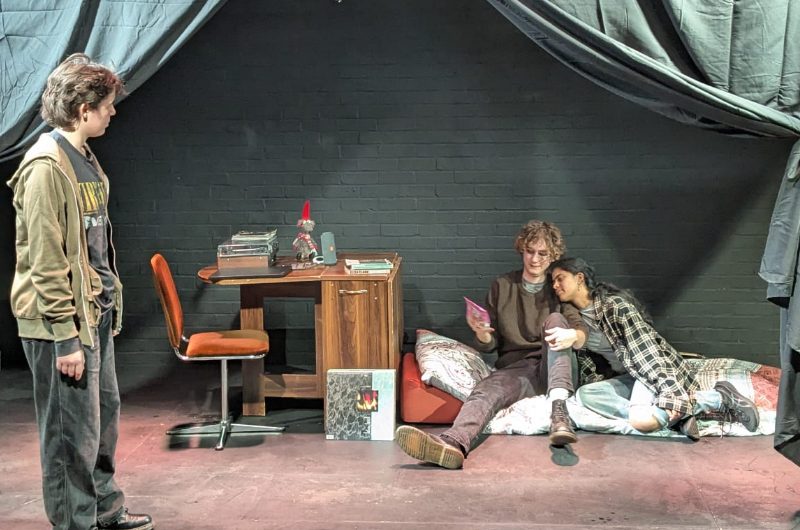 A photo of three cast members on set. Two are lying down whilst one is standing and speaking to them.