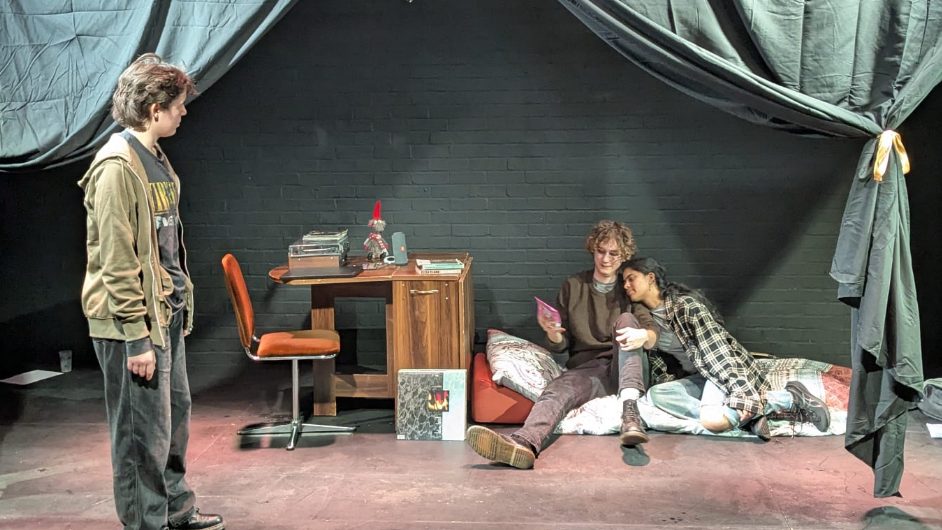 A photo of three cast members on set. Two are lying down whilst one is standing and speaking to them.