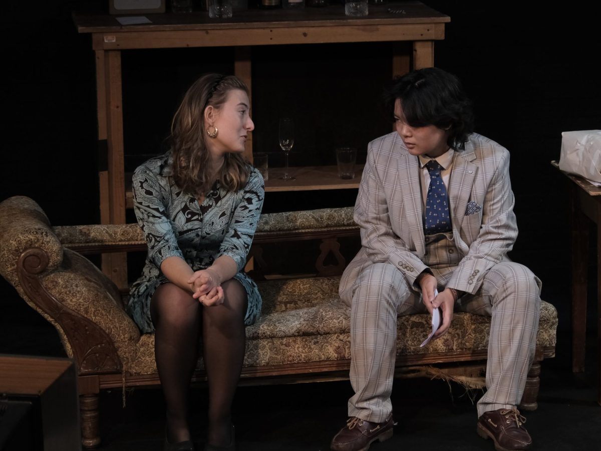 An image of two cast members sat on a sofa of the set looking at each other