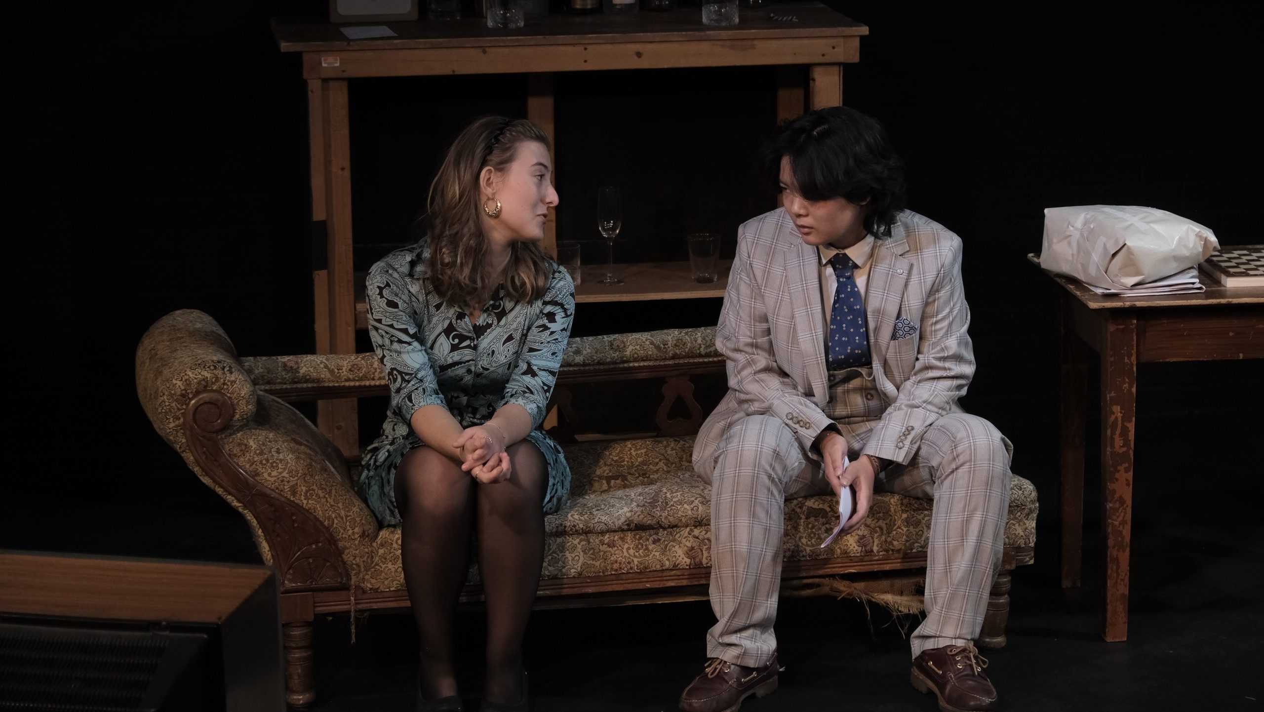 An image of two cast members sat on a sofa of the set looking at each other