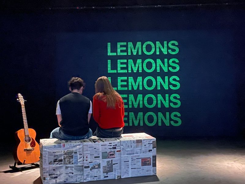 Two actors sit on a bench, next to a guitar.