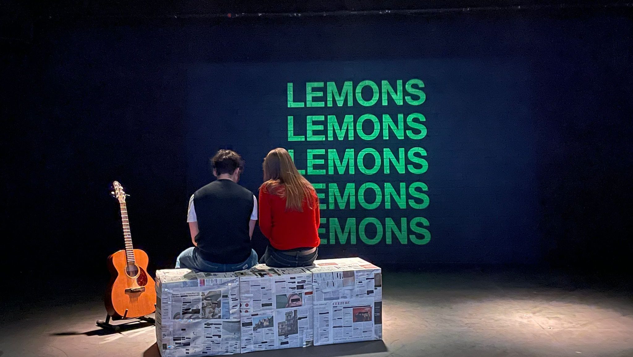 Two actors sit on a bench, next to a guitar.