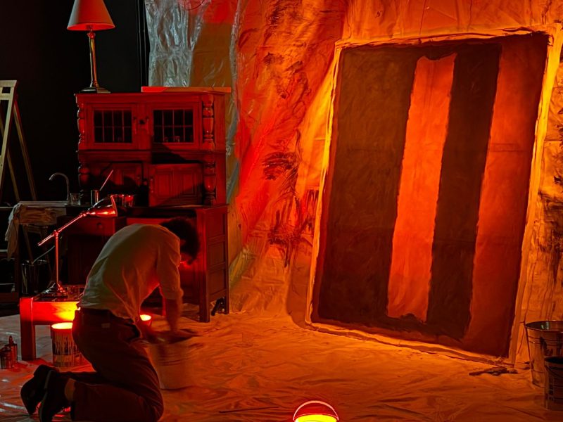 A man kneels on the ground in front of a large painting.