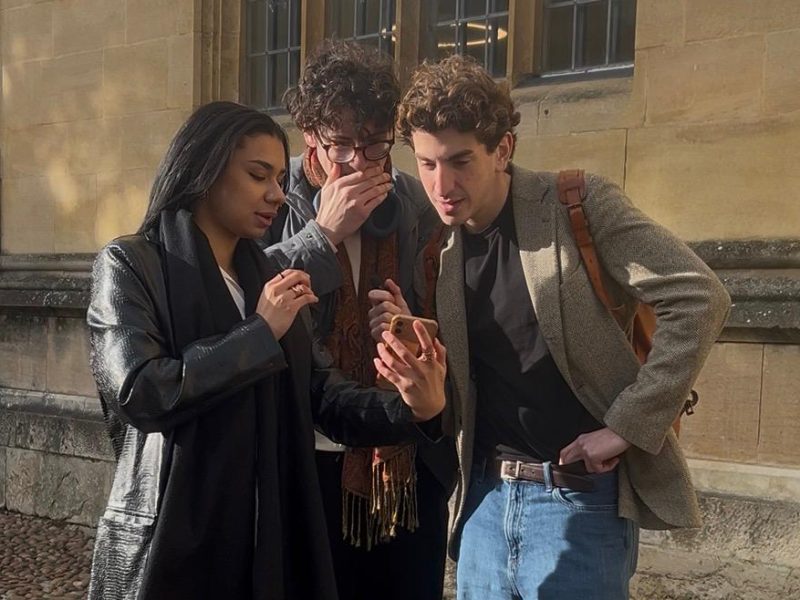 Three people look at a phone. They seem shocked.