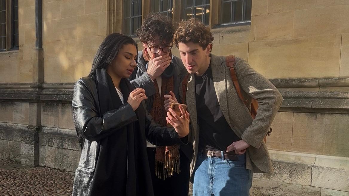 Three people look at a phone. They seem shocked.