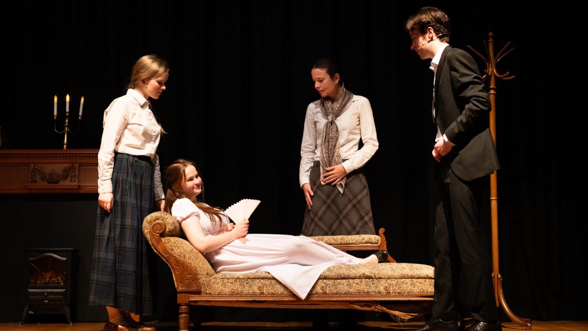 A group of people stand around a woman lying on a chaise longue.