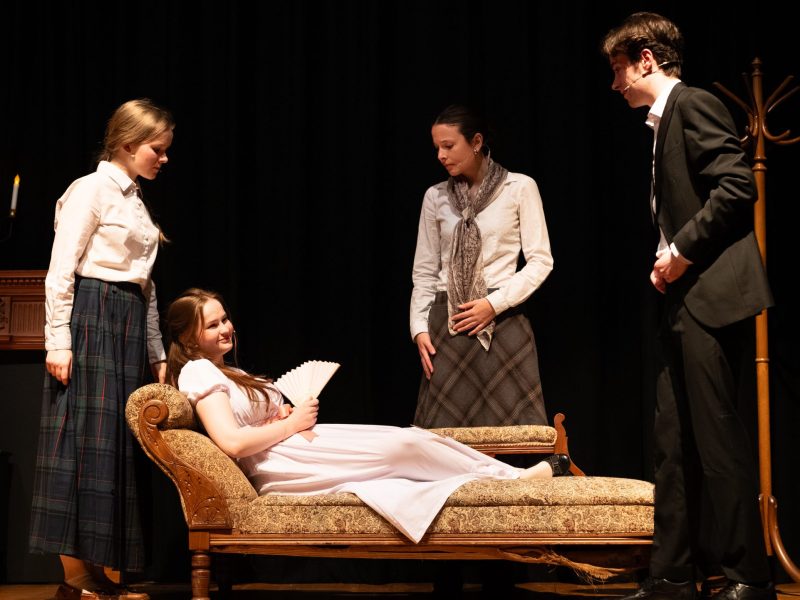 A group of people stand around a woman lying on a chaise longue.