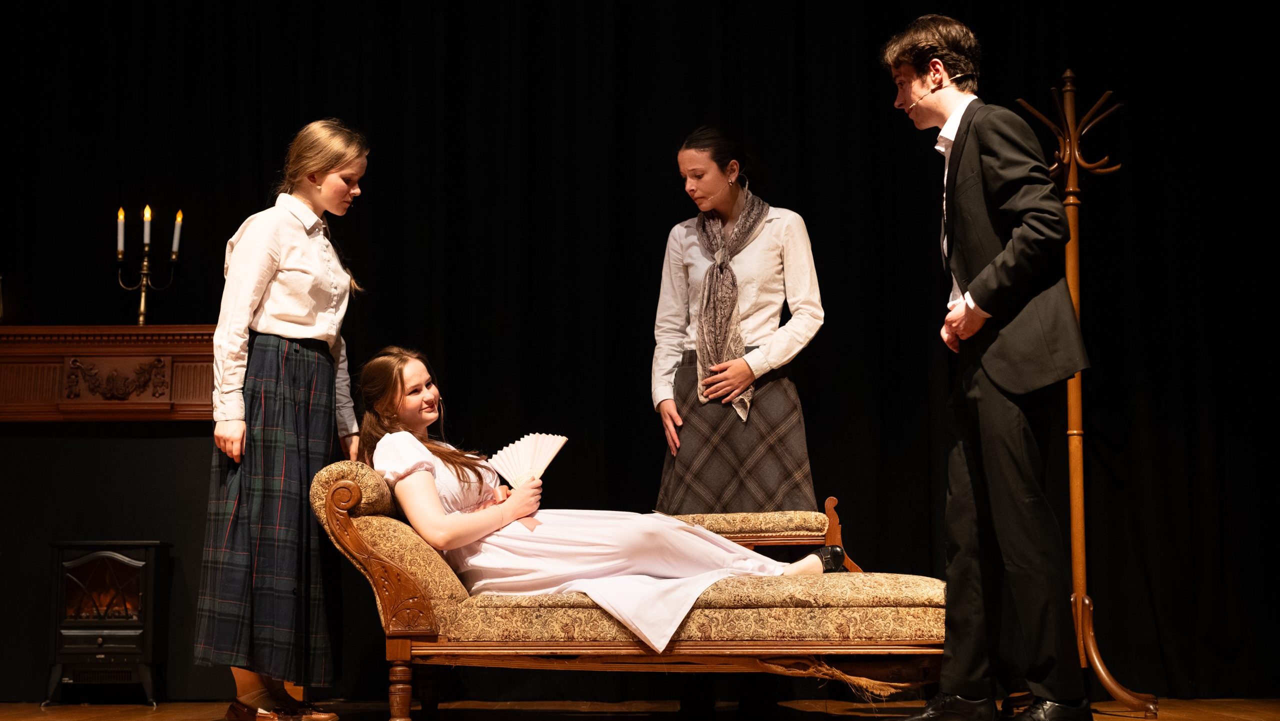A group of people stand around a woman lying on a chaise longue.