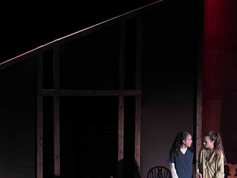 A student theatre set with two actors and a wooden structure which looks like a house.