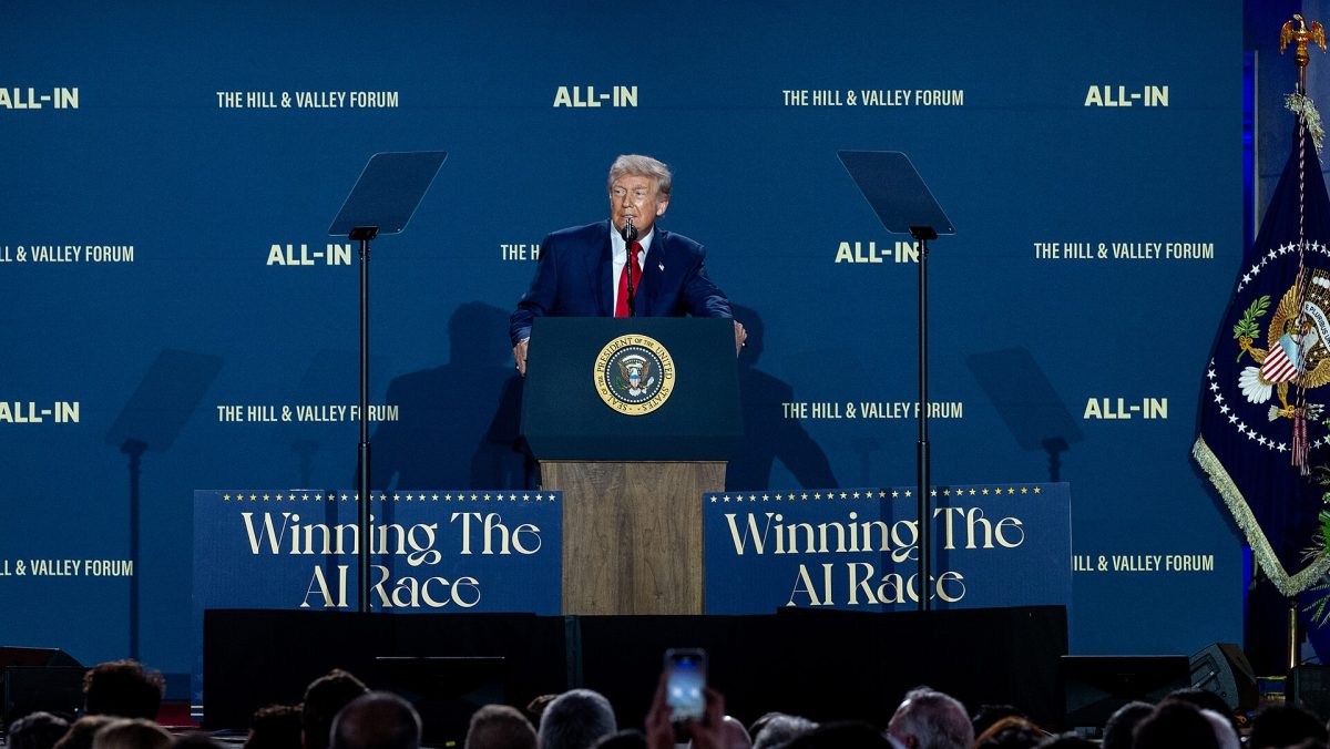 President of the United States Donald Trump stands at a podium with large blue signs on both side saying "Winning the AI Race".