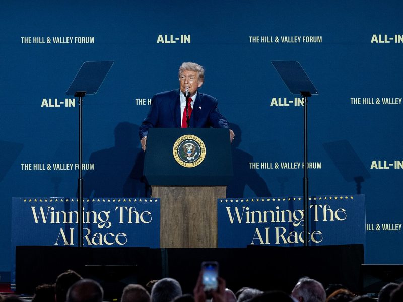 President of the United States Donald Trump stands at a podium with large blue signs on both side saying "Winning the AI Race".