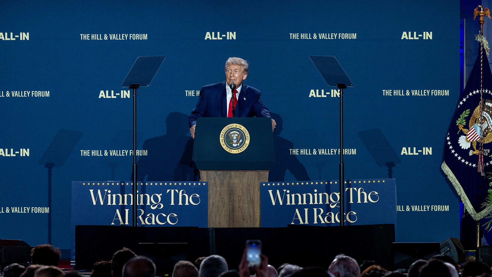President of the United States Donald Trump stands at a podium with large blue signs on both side saying "Winning the AI Race".