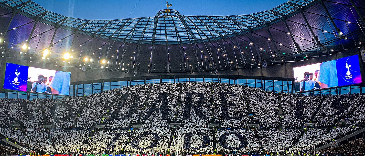 The Tottenham Hotspur Stadium, pictured in the evening. A card mosaic tifo reads "to dare is to do."