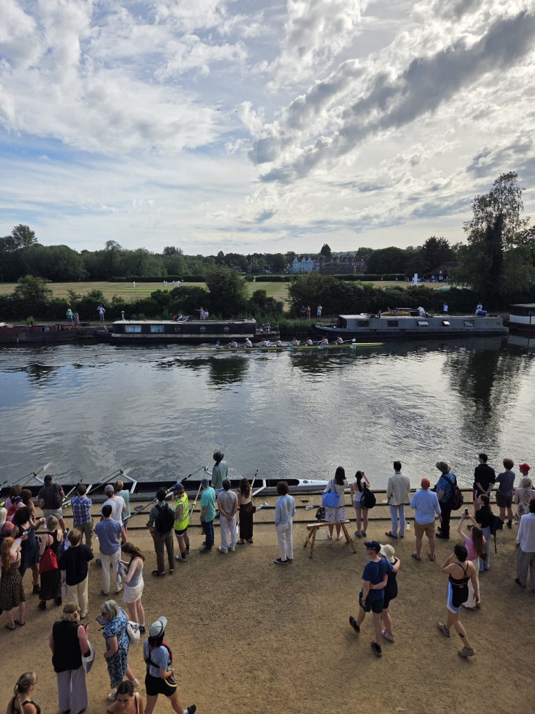 The Oriel M1 boat races to the finish as cricketers play in the background.