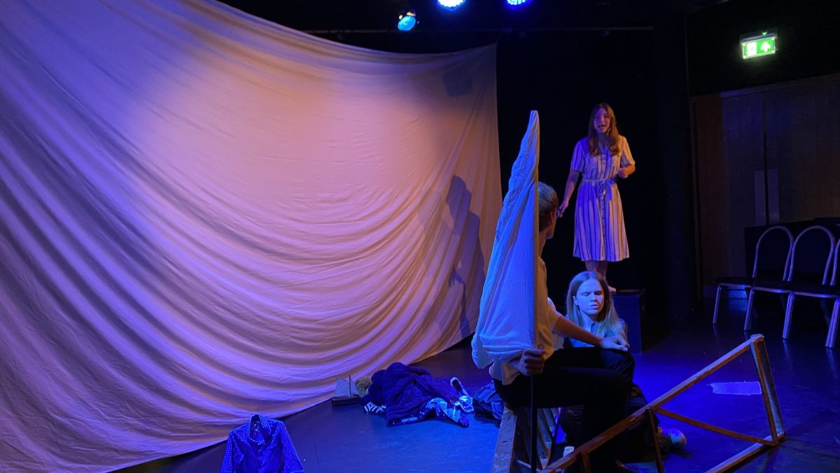 Four actors gesticulate to each other in front of a large white sheet. The photo is taken at a side angle.
