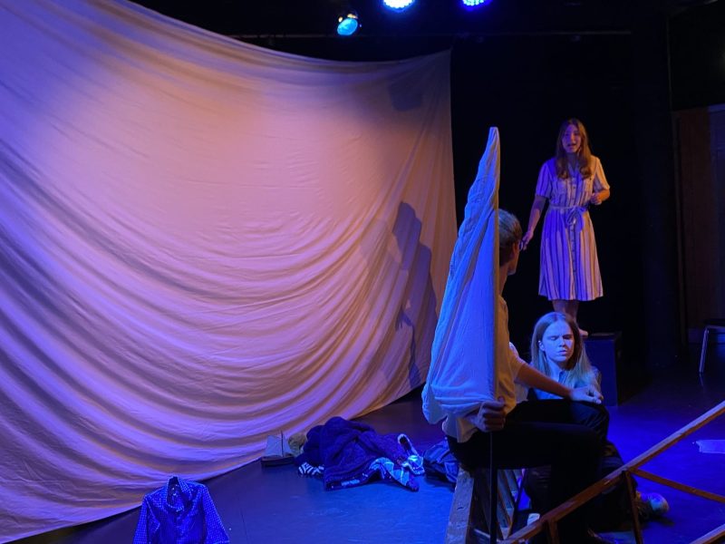 Four actors gesticulate to each other in front of a large white sheet. The photo is taken at a side angle.