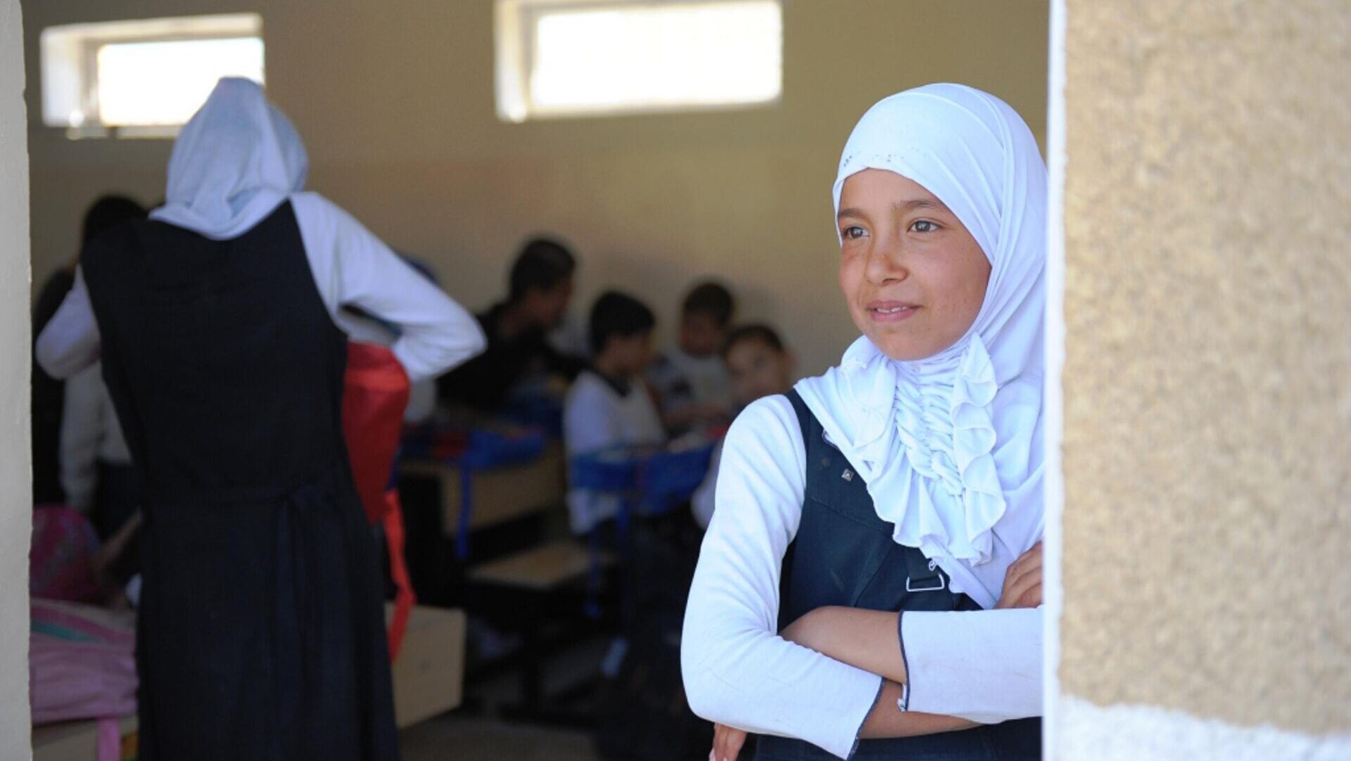 A girlin a hijab stares out of her classroom - behind her a teacher is teaching students