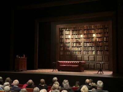 Image of Oxford Playhouse's 'The Gang of Three' set, consisting of a large bookshelf, sofa, and the audience below the stage.