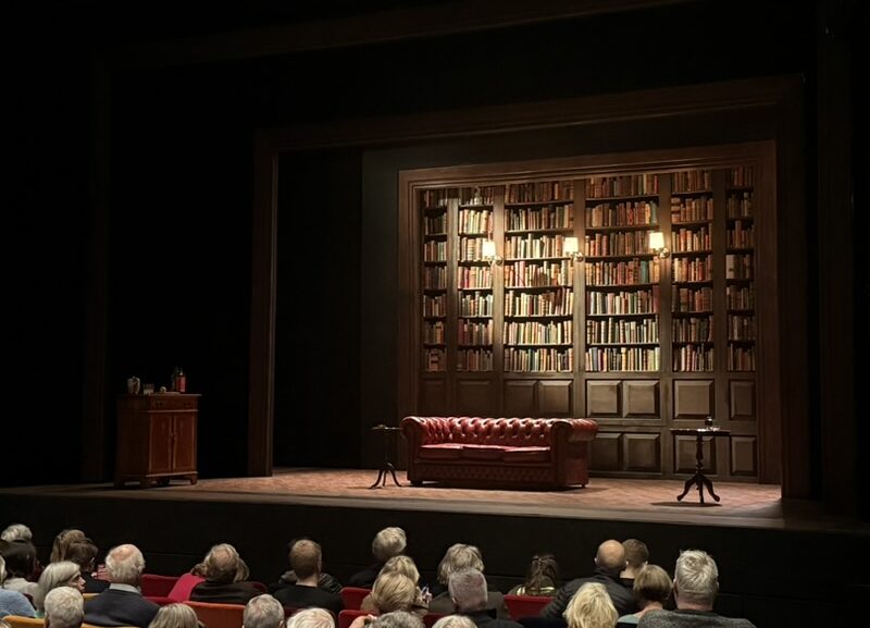 Image of Oxford Playhouse's 'The Gang of Three' set, consisting of a large bookshelf, sofa, and the audience below the stage.