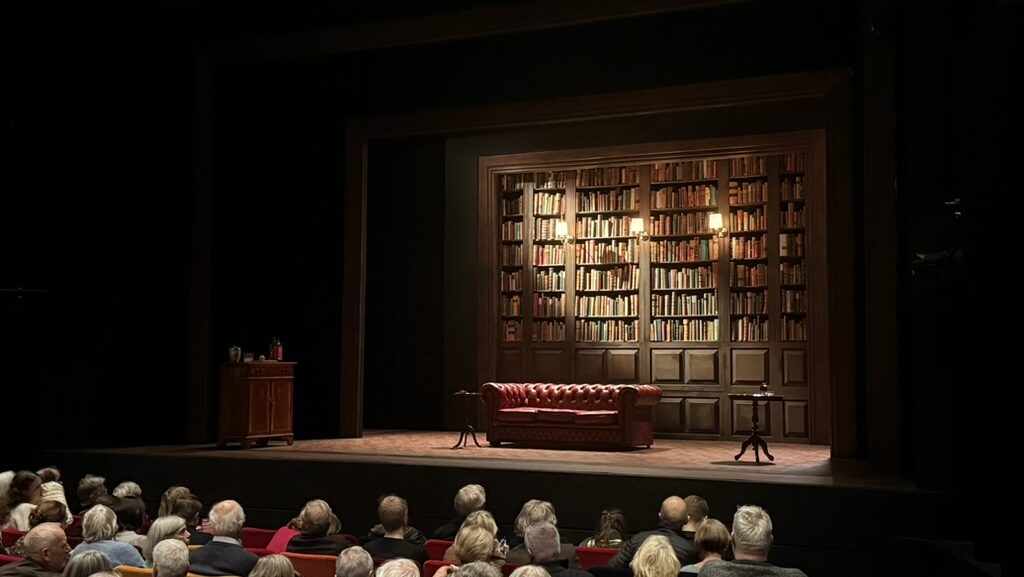 Image of Oxford Playhouse's 'The Gang of Three' set, consisting of a large bookshelf, sofa, and the audience below the stage.