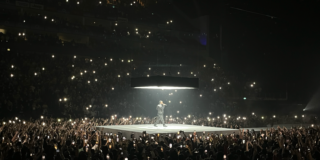 A photograph of Kendrick Lamar performing in London. Kendrick Lamar stands on the stage, surrounded by a crowd waving lights in the air.