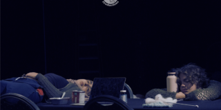 Dim lighting, two people lying on top of a table next to a laptop and various items as part of The Antipodes's setting