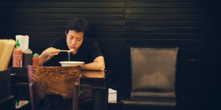 A man sits at a table alone, eating a bowl of ramen.