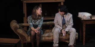 An image of two cast members sat on a sofa of the set looking at each other